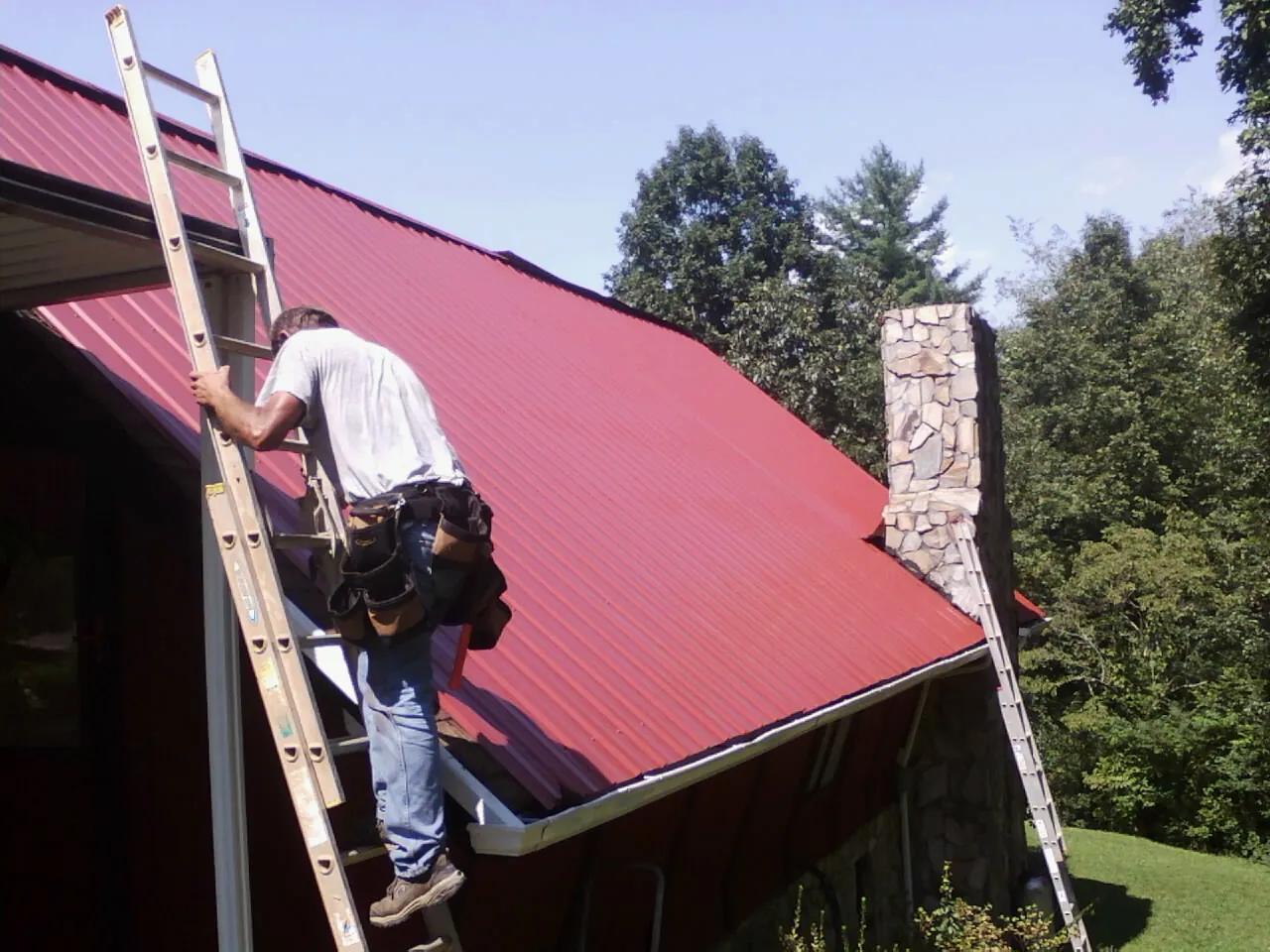 Roofer on a ladder beside a red metal roof and stone chimney — Soffit & Fascia Repair by Roofing Brothers in Marstons Mills