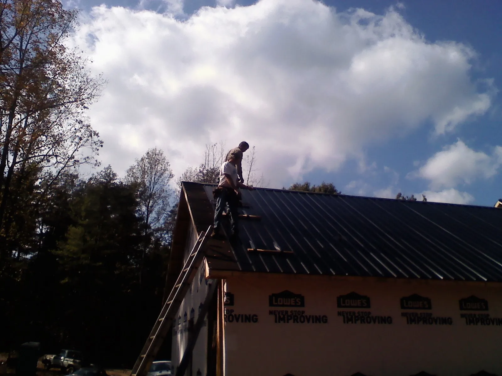 Roofer fastening dark metal panels high against a cloudy sky, from a Roof Inspection job in Marstons Mills
